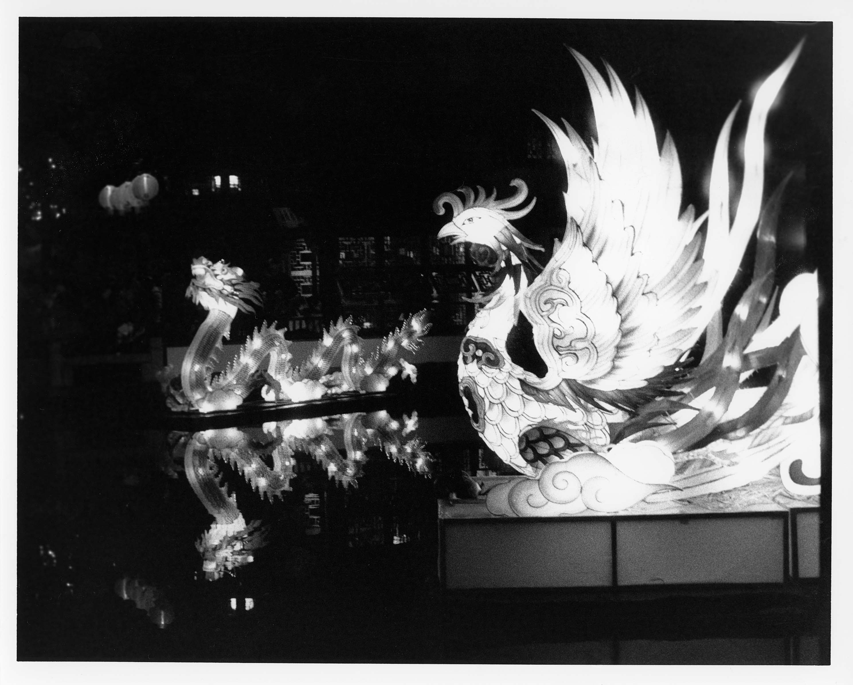 Black and white photo of a dragon and a phoenix statue floating in water. The reflections perfectly mirror the dragon. It is night